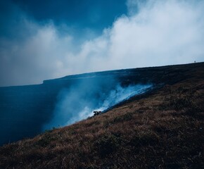 Dramatic volcanic smoke plumes rise from a barren, grassy mountainside under a cloudy sky