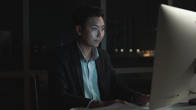 Asian businessman working late night at office desk in dark modern corporate environment with city lights in background, focused professional using desktop computer overtime after hours.