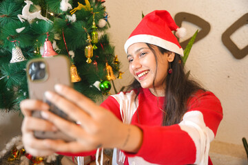 Young indian woman wearing santa hat taking selfie picture with smart phone and celebrating Christmas in front of a decorated tree at home.