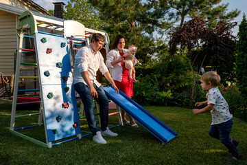 Fototapeta premium Parents crouch by a blue slide holding their baby as a toddler runs toward them, colorful playground set and green garden creating an active home scene