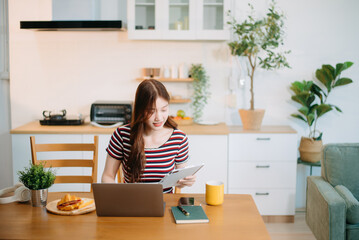 Young woman enjoying coffee and pastries while working remotely from her cozy kitchen on laptop and tablet, embracing a relaxed