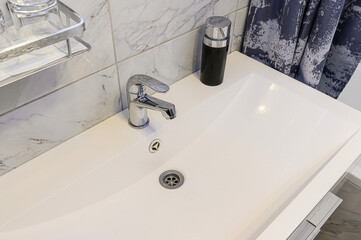 Close-up of modern white porcelain sink with chrome single-lever faucet and pop-up drain. Black matte soap dispenser, silver wire shelf holding glasses, gray marble-effect tiles