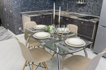A modern kitchen scene featuring a glass dining table set with dinnerware, candles, and champagne glasses, surrounded by minimalist chairs