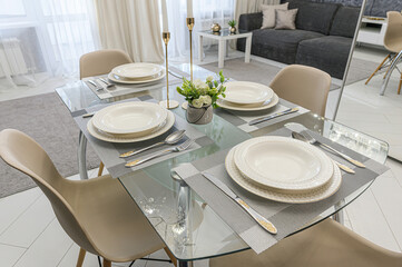 A dining area features a glass table set for four, complete with plates, cutlery, and a small floral arrangement. Minimalist décor complements the bright, neutral tones