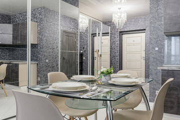 A modern dining area with a glass table set for a meal, surrounded by beige chairs. Mirrored walls reflect the kitchen and doors, enhancing the room's spacious feel