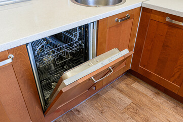 An open dishwasher is nestled in wooden cabinets, showcasing the metal racks. A light countertop...
