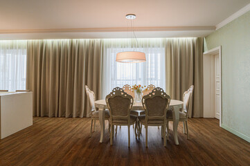 Elegant dining room: round white table with ornate beige-upholstered chairs, yellow daisy vase centerpiece, large beige curtains on bay windows, white drum pendant, mint floral walls, oak floor