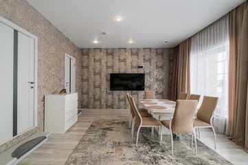 A dining room features patterned wallpaper, a white dining table surrounded by beige chairs, and a TV mounted on the wall. Neutral curtains frame a window