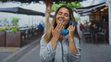Woman holding blue telephone receiver, hand covering mouth at street cafe terrace near outdoor seating; surprise friendly conversation. - Powered by Adobe