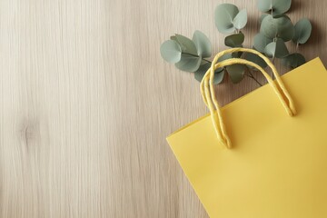 A vibrant yellow shopping bag rests on a light beige wooden surface, accented by sprigs of soft green eucalyptus.