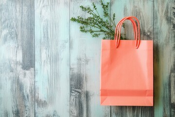 A coral-colored paper shopping bag sits atop a distressed wooden surface, with sprigs of greenery for a spring-like aesthetic.