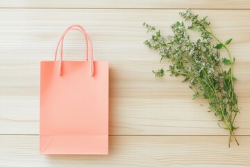 A pale peach gift bag sits atop a light beige wooden surface, complemented by a bouquet of delicate white-flowered herbs.