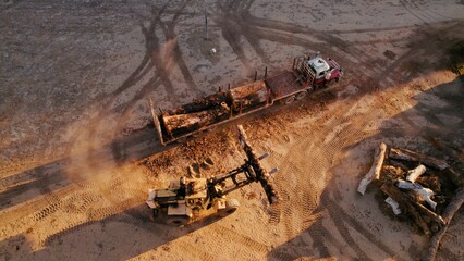 Heavy machinery operates on a construction site in a rural area at sunset, moving logs and preparing the landscape