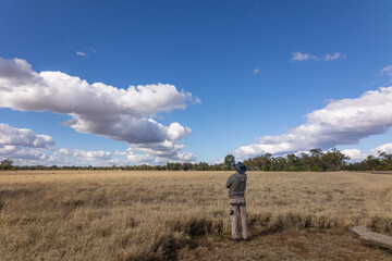 A farmer stands and looks hopefully over a field of Mitchell grass and a line of gumtrees at some interestingly shaped clouds in a dry region at Charleville in outback Queensland, Australia.