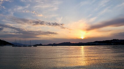 Naklejka premium Sun dips below the hills, painting the sky with warm hues as it reflects off the gentle sea, with sailboats silhouetted on the tranquil water.