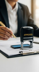 Close-up of gold 'APPROVED' stamp on document with businessperson. Professional office setting with clipboard and pen. Corporate approval process in a work environment.
