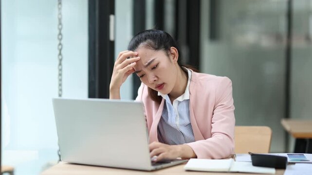 Professional Asian businesswoman suffering workplace stress, holding head while typing on laptop, looking overwhelmed in corporate office setting