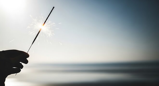 Silhouette of a hand holding a lit sparkler against a hazy beach sunset