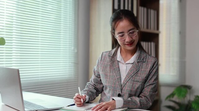 Professional businesswoman wearing glasses, typing on laptop and writing notes at workspace with concentrated demeanor