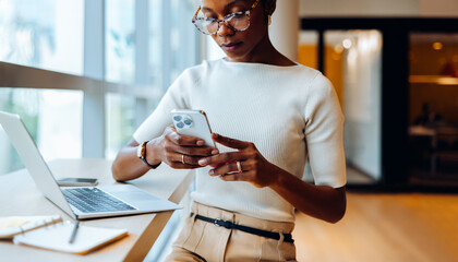 Fototapeta na wymiar Businesswoman reading her smartphone while working on her laptop at a desk