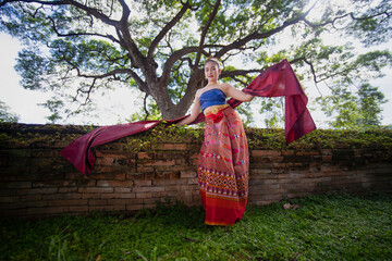 Woman in traditional attire poses gracefully with red scarf in front of ancient brick wall and large tree