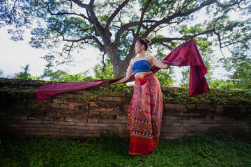 Traditional Thai dancer in colorful attire poses gracefully by ancient brick wall under large tree