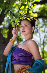 Traditional attire woman under tree, wearing gold jewelry, looking thoughtful. Sunlight filters through leaves, creating serene atmosphere