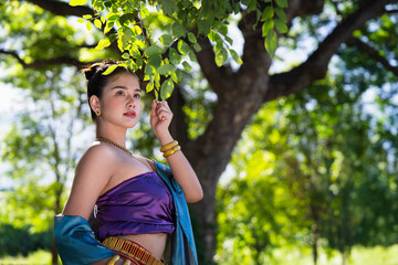 Woman in traditional attire stands gracefully under tree, surrounded by lush greenery and sunlight