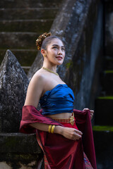 Woman in traditional attire stands gracefully against stone staircase, wearing blue top and red wrap, adorned with gold jewelry