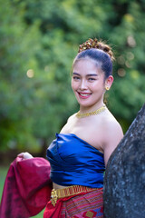 Woman in traditional Thai attire smiles warmly, wearing blue top and red patterned skirt, with gold jewelry, set against lush green background