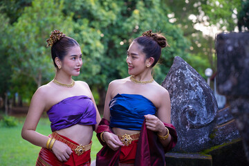 Two women traditional Thai attire stand outdoors, wearing colorful silk tops and red skirts, adorned with gold jewelry. They are smiling