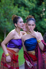Traditional Thai attire with two women sharing secret, wearing colorful silk tops and patterned skirts, adorned with gold jewelry, lush garden