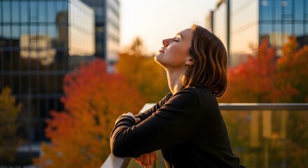 Young woman on a sunny city balcony, with eyes closed and face turned to the sun, enjoying a serene moment amidst autumn trees and modern buildings.