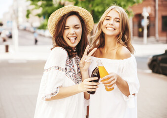 Two young stylish smiling hippie brunette and blond women models in summer sunny day in white hipster clothes posing on the street background. Drinking soda from bottle
