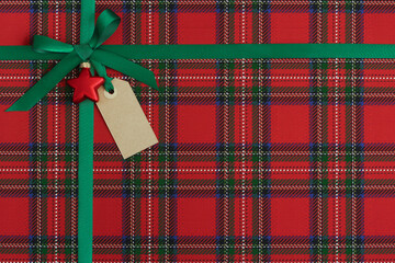 Christmas present, gift close-up. Green satin ribbon, bow with red ornament and empty tag on plaid paper background.