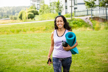 Middle aged woman of color walking with yoga mat to park, embracing diversity, wellness, and active modern lifestyle in nature.