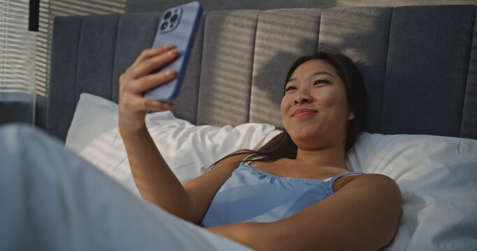 Girl Having Morning Video Call. Young Asian Woman Lies in Bed With Smile, Holding Smartphone During Video Call. Concept of Remote Communication, Online Connection, Morning Routine, Digital Lifestyle.