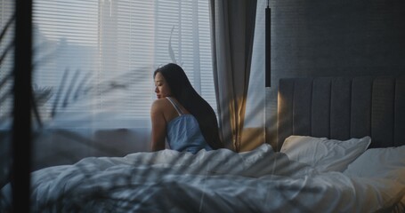 Asian Woman in Nightwear Sits on Bed By Window in Soft Morning Light. Girl Wakes Up in Bed, in...