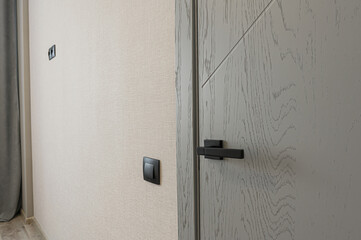 Close-up of modern interior featuring grey door with sleek black handle, complemented by black light switch on textured cream-colored wall. Light wood flooring adds to the room contemporary aesthetic