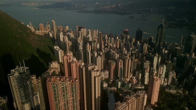 Steep hillside meets dense towers as Victoria harbour opens beyond. Evening sun paints tall blocks and busy water routes while ferries trace lines and hazy ridges frame Hong Kong panorama