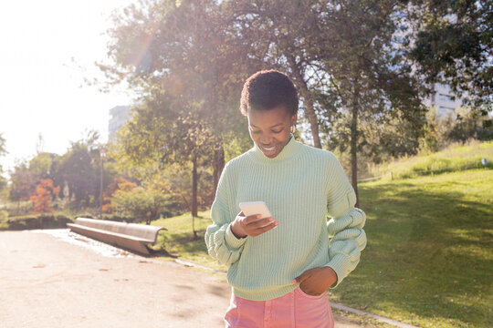 young african american woman smiling happy while using cell phone, she is standing in a park with trees in the background, concept of technology of communication, copyspace for text - Powered by Adobe