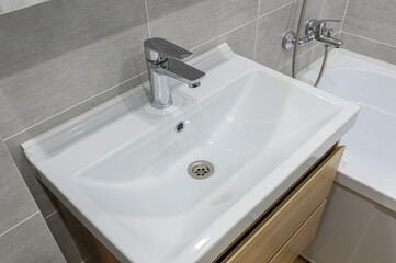 A clean bathroom sink is shown, featuring a chrome faucet. The sink is white with a wooden cabinet underneath, set against a backdrop of gray tiles