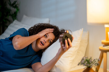 Latina young woman holding alarm clock while lying down on bed at night. 
