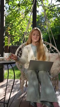 Middle aged woman working remotely outdoors on terrace with laptop and coffee mug. Cozy lifestyle, natural light, peaceful atmosphere, balance between work and life.