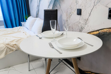 A round white table is set for two with plates, silverware, and wine glasses in a modern room. The background features a bed and a decorative wall