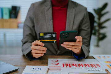 Asian businesswoman using smartphone and smiling while working with documents and computer at office desk, concept of modern technology and business administration