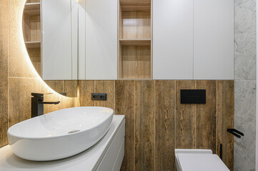 Modern bathroom featuring an oval white sink, black faucet, wood-paneled wall, illuminated round mirror, and sleek cabinets