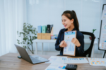 Young Asian businesswoman holding a pen and clipboard, analyzing statistical data on a tablet screen while working on a computer in a modern office environment