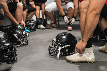 American football players getting ready in a locker room, putting on cleats and tying laces, with helmets on floor