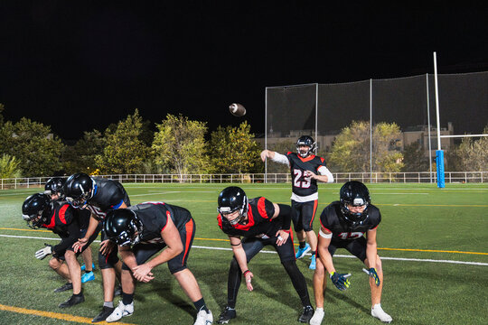 American football players practicing on a lighted stadium field at night, a quarterback throwing the ball to teammates